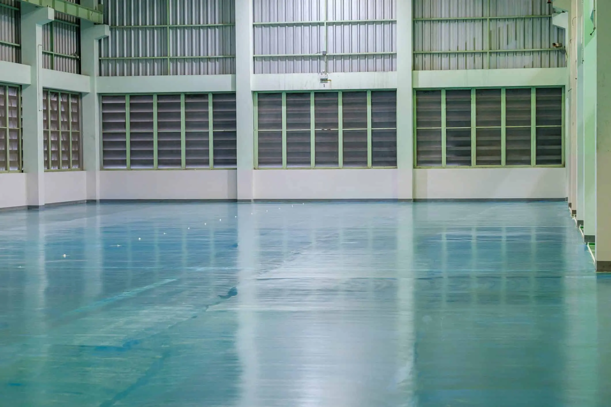 Empty industrial warehouse with a shiny blue floor, white walls, and large shaded windows on one side.
