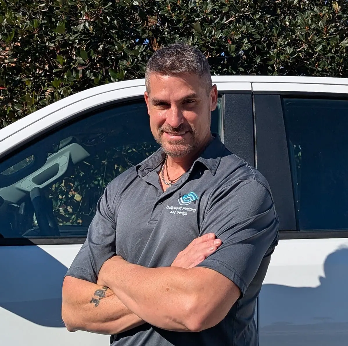 Man with short hair and beard stands in front of a white vehicle, arms crossed, wearing a gray polo shirt with a small logo, with greenery in the background.