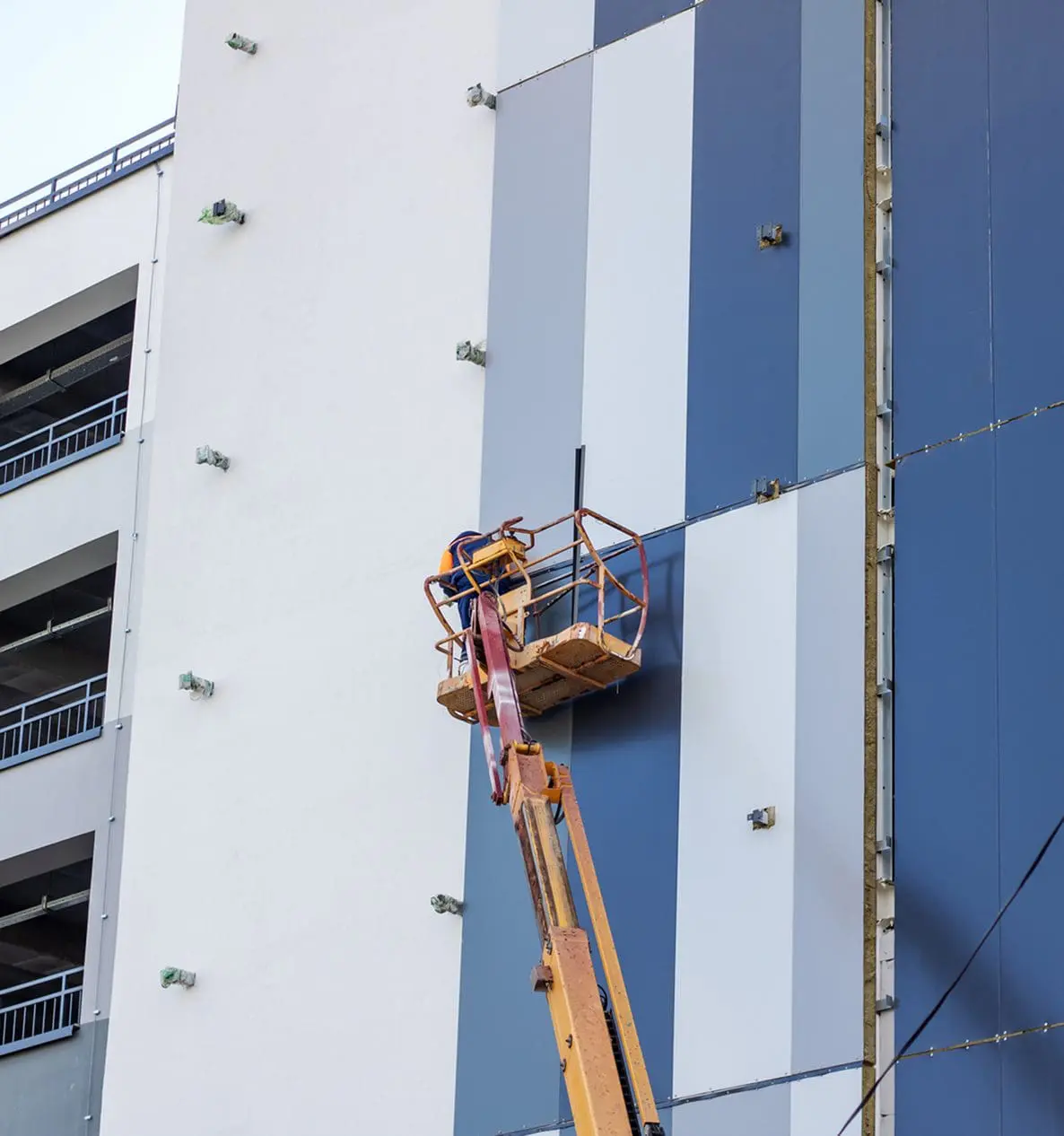 A worker operates a boom lift to install or maintain panels on the exterior of a tall, striped building.
