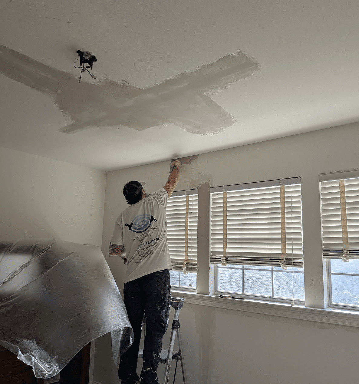 Person painting ceiling near covered furniture.