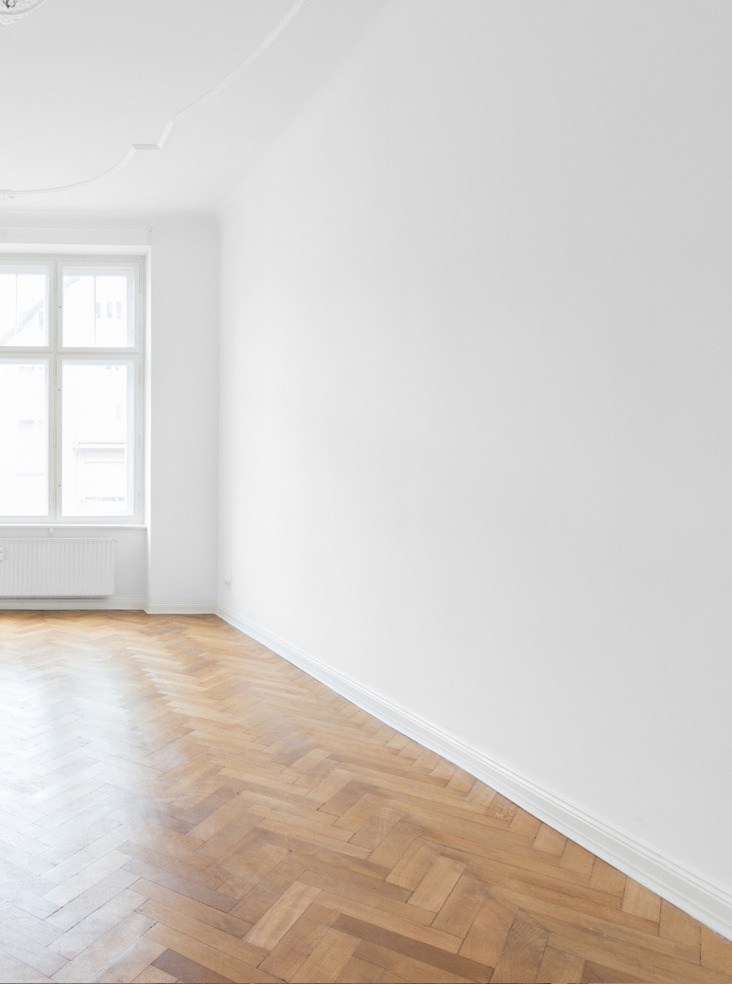 Empty room with white walls, a large window, and light wooden herringbone-patterned floor. Natural light enters from the window on the left side of the image.