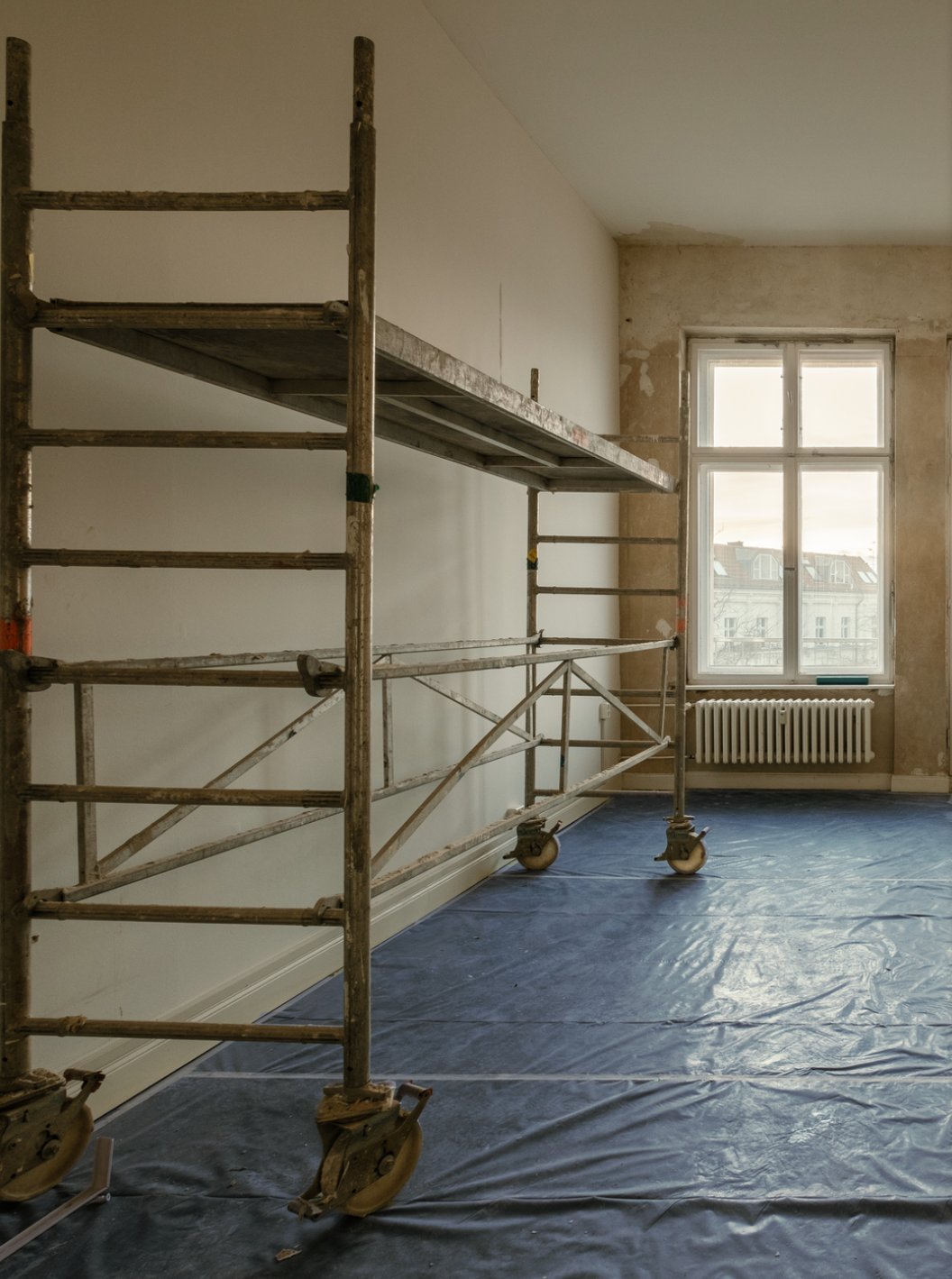Metal scaffolding stands on a blue plastic-covered floor in a partially renovated room with a large window and exposed radiator.
